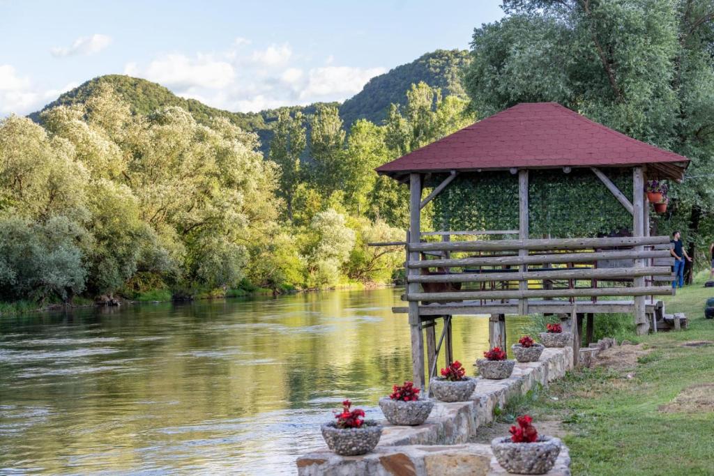 Überdachter Holzpavillon direkt am Flussufer, umgeben von grünen Hügeln und mit roten Blumen geschmückten Steintöpfen — reale Ferienunterkunft am Balkan, die erfolgreich über Airbnb und Booking.com vermietet wird